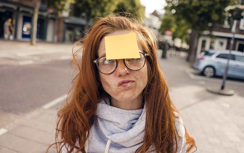 A red-haired white woman wearing glasses stands on a street, grimacing at a yellow sticky note stuck to her forehead