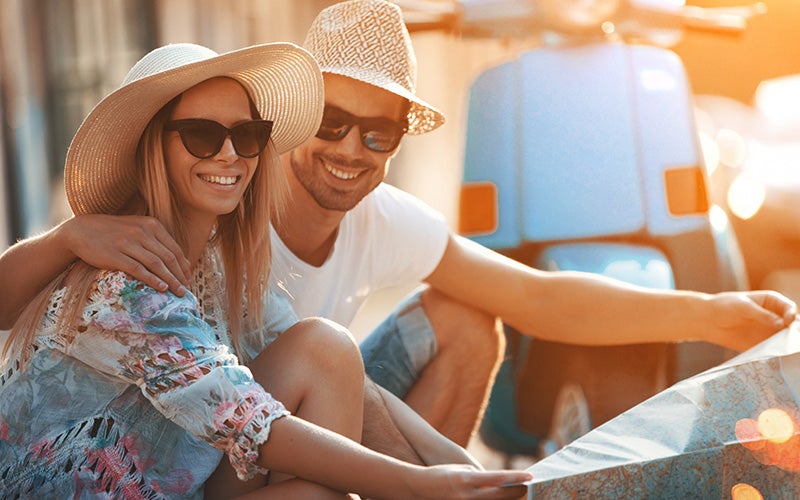 Una coppia seduta sul marciapiede con cappelli e occhiali da sole studia una mappa
