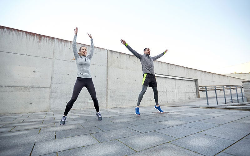 A man and a woman in long-sleeved sports gear do jumping jacks outdoors on large paving stones. 