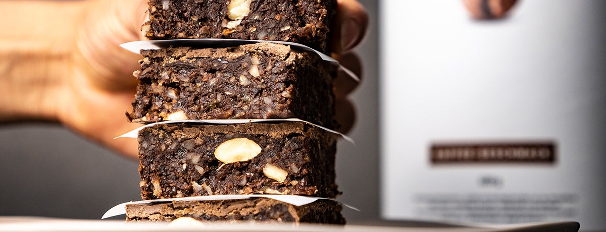 A medium-skinned hand in the background reaches for the top brownie on a stack of raw vegan coffee brownies, packed with chunks of nuts and deliciousness