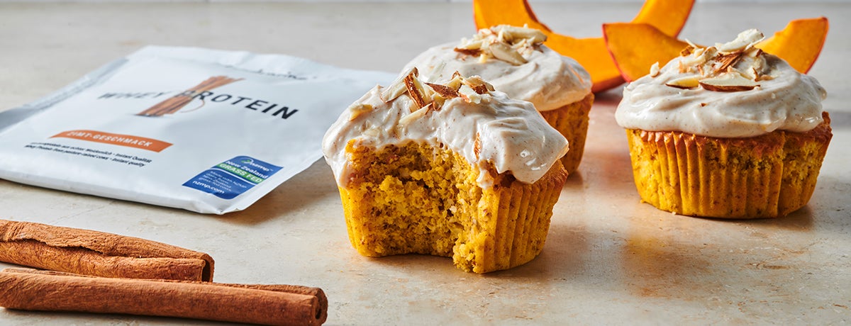 Two protein pumpkin muffins topped with creamy frosting, with a package of Whey Protein and slices of bright orange sunshine squash in the background.