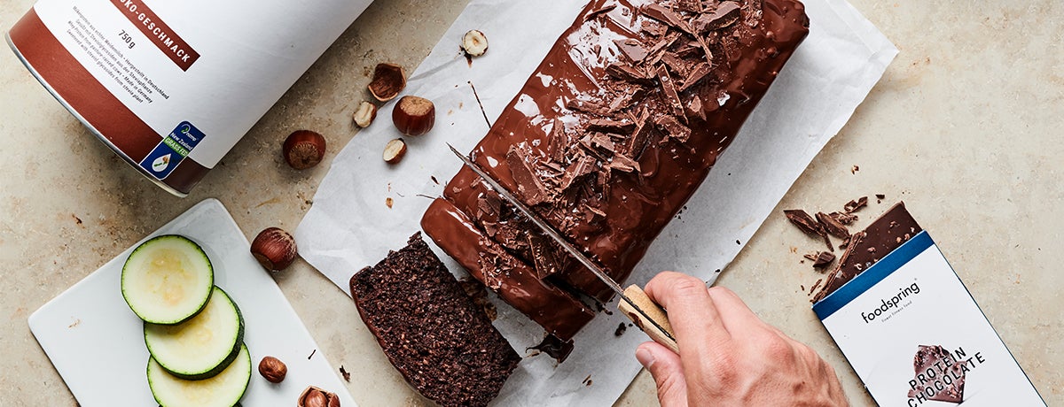 A hand holding a knife cuts a slice off of a low-carb chocolate zucchini cake drizzled with protein chocolate ganache.