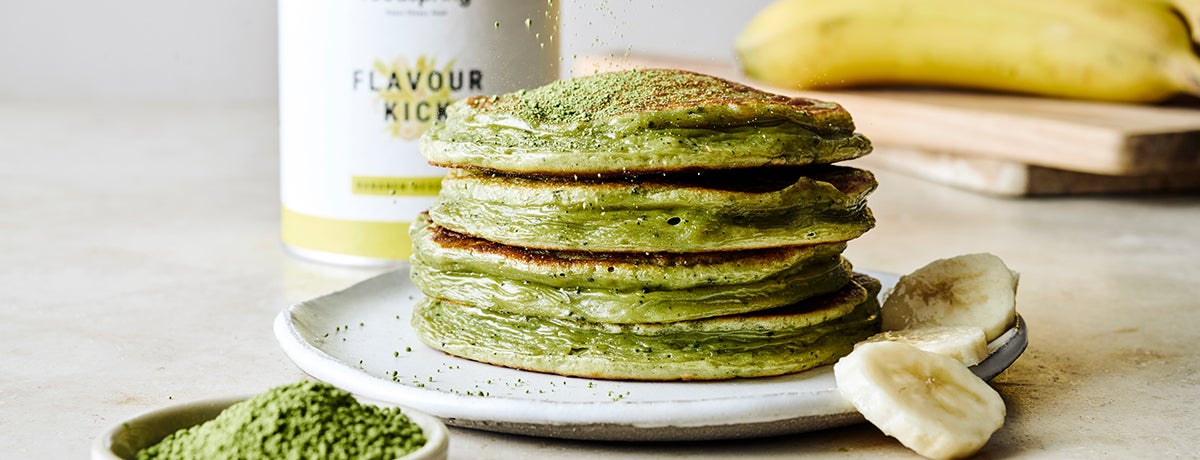 A stack of green Matcha Pancakes with a banana and a canister of Banana Flavor Kick in the background