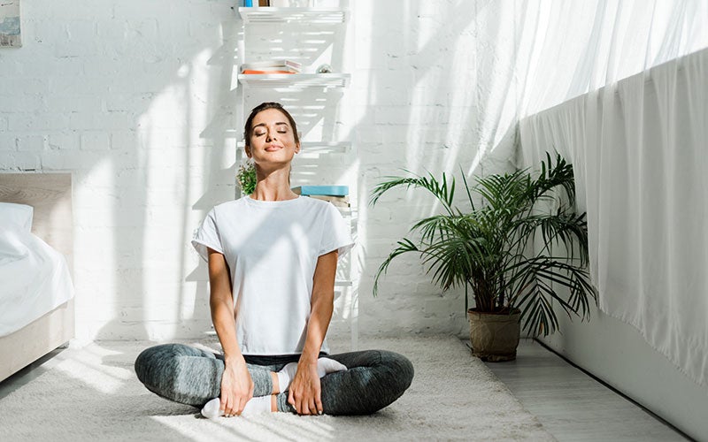 A white woman sits indoors in a white-walled bedroom with her eyes closed practicing mindfulness through meditation