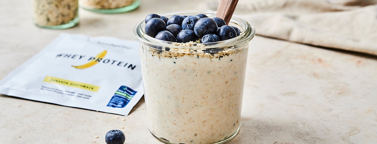 a glass jar of overnight oats topped with fresh blueberries sits next to a single-serving package of foodspring's banana Whey Protein