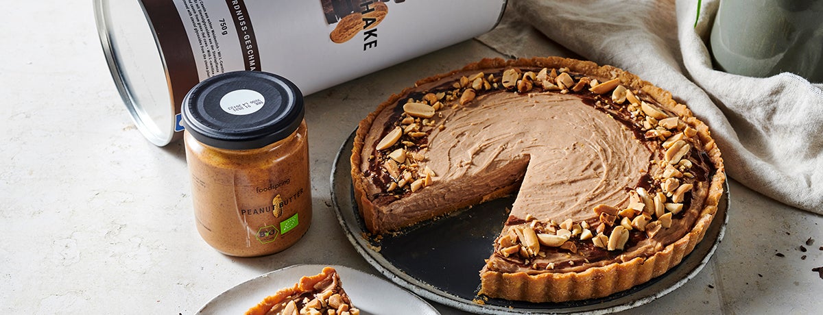 a rich brown chocolate peanut butter cheesecake, with a generous slice cut out of it, sits on a dark gray serving dish. The rim of the cheesecake is lined with crushed peanuts. A jar of foodspring Peanut Butter sits next to the cheesecake.
