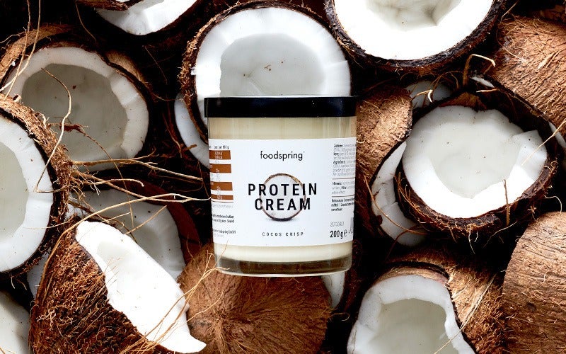 a jar of foodspring's Coconut Crisp Protein Cream on a background of halved coconuts