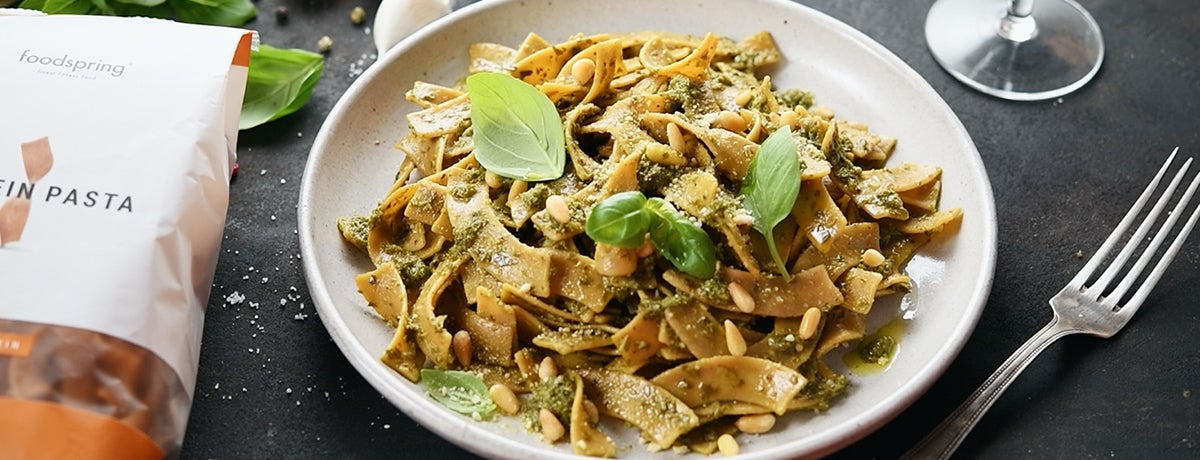 A plate of Protein Pasta with a lemon balm pesto, basil leaves, and pine nuts on an anthracite-gray table