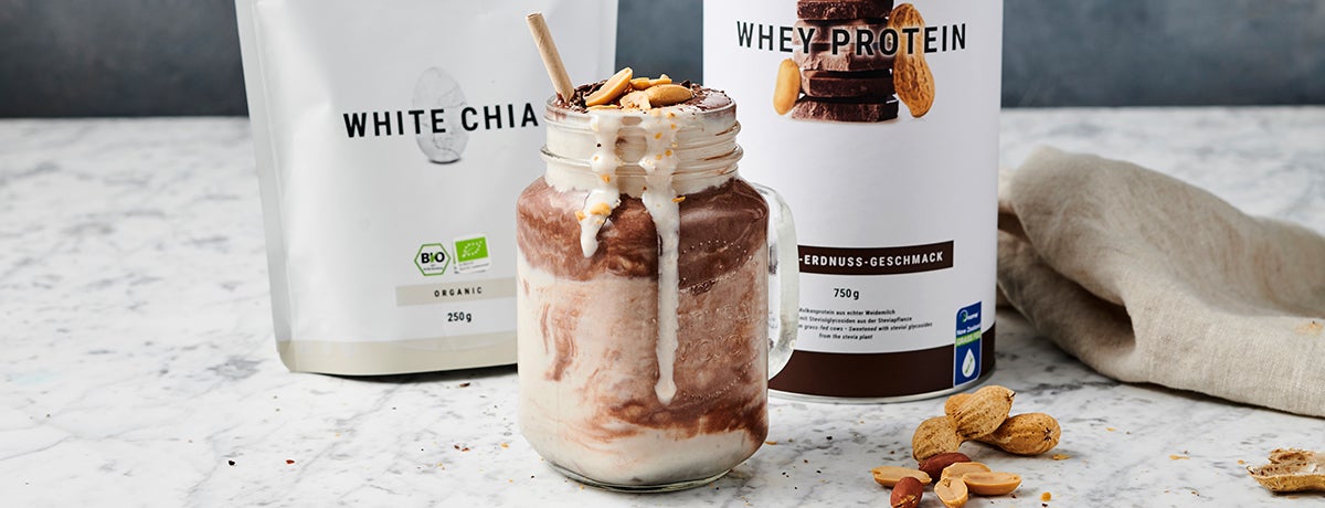 A mason jar filled with layered chocolate-peanut butter protein shake, with some whey protein dripping decoratively down the side, toppede with peanuts, a paper straw sticking out, and displaying White Chia Seeds and Chocolate Peanut Butter Whey Protein in the background.
