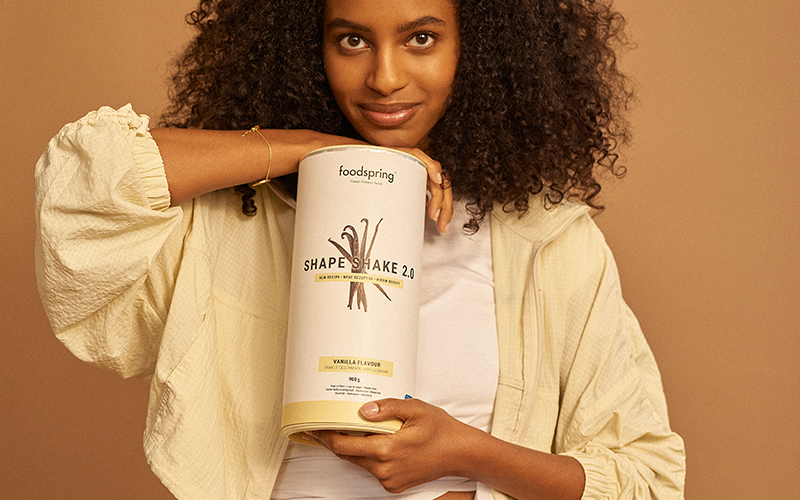 A woman of color with a small smile and curly dark-brown hair holds a canister of Shape Shake 2.0 meal replacement shakes in front of a mocha-colored background.