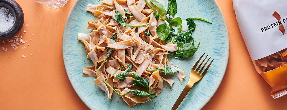 A plate of Protein Pasta with a creamy cheese sauce garnished with green spinach and with a golden fork laid next to it