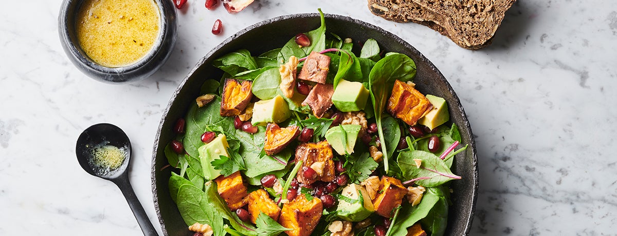 An anthracite-gray plate holds a plate of deep green salad leaves with oven-roasted sweet potato cubes on top. It is garnished with deep red pomegranate seeds and bright yellow-green avocado chunks. There is a bowl of mustard-yellow dressing on the side.