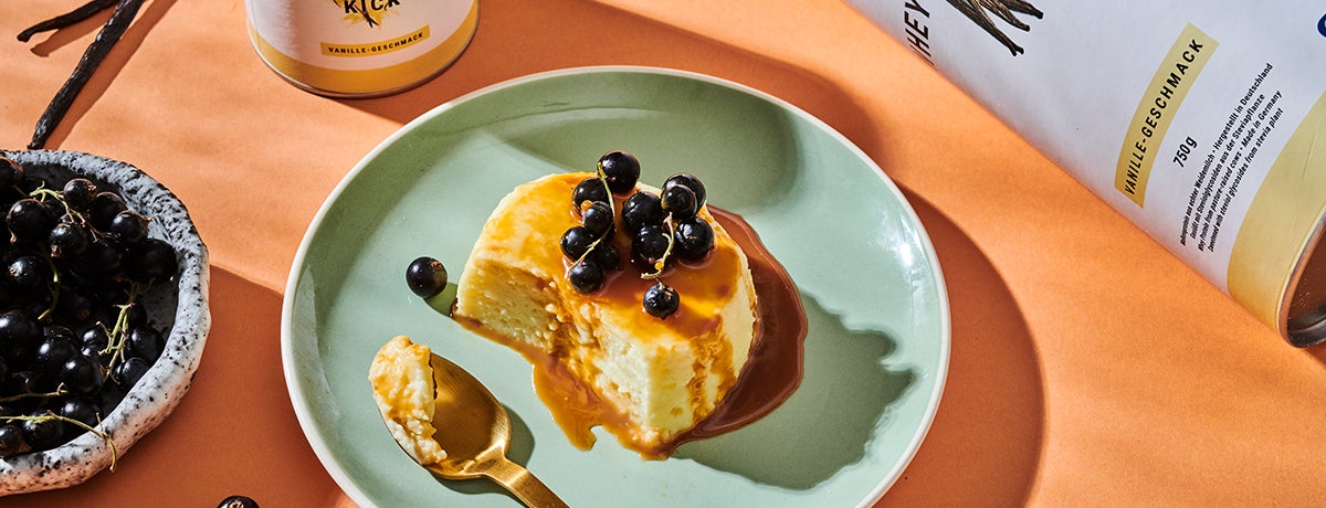 a photo of a vanilla protein flan, drizzled with caramel and topped with blueberries, on a sea-green plate sitting on a coral-colored tablecloth