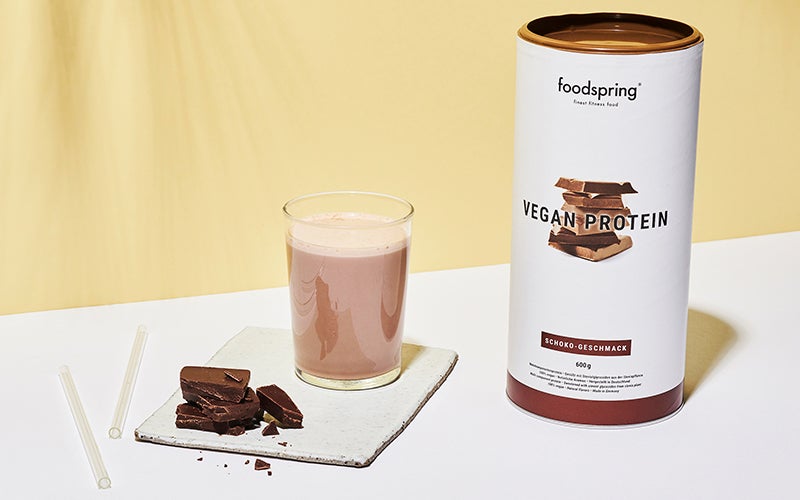 A canister of foodspring's chocolate Vegan Protein next to a glass full of protein shake.