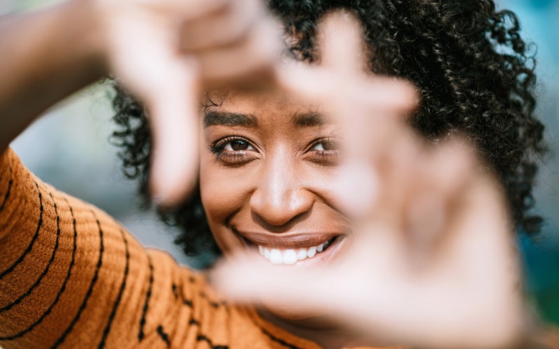 A woman of color looks through a square formed by her fingers, giving a big smile to the camera.