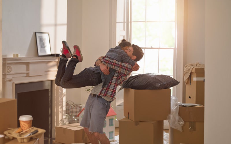 A man and woman hug in an unfurnished room full of boxes. The man is lifting the woman into the air and her feet are fully off the ground behind her.