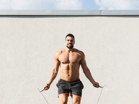 a shirtless, muscular man performs a jump rope workout outdoors in front of a cream-colored wall