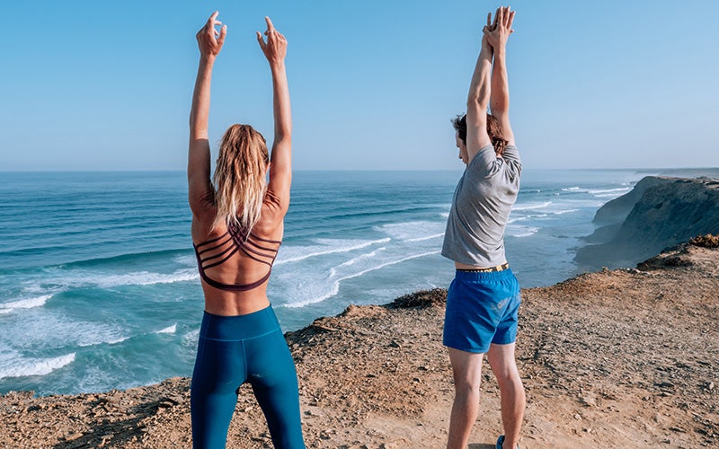 Two athletes stretch at the top of a cliff. They are seen from behind with the ocean in front of them.