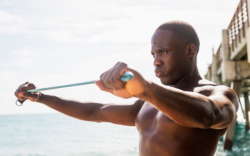 A shirtless man of color puffs his cheeks out to exhale while doing a shoulder exercise with a resistance band. He stands outdoors with the ocean and a pier behind him.