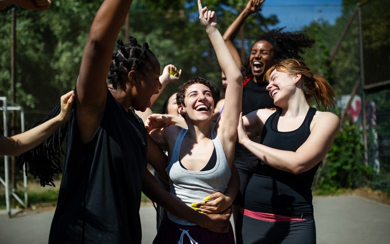 A group of women of many races, hair colors and lengths smile and seem to celebrate together on an outdoor basketball court