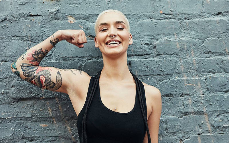 A white, tattooed woman with a bleached buzz cut grins in front of a wall while making a strong biceps gesture to show off her lack of bingo wings