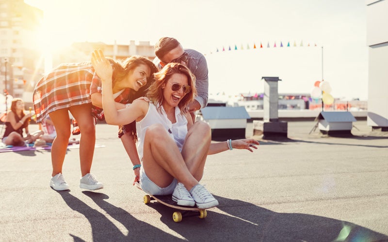 Una donna siede su uno skateboard e viene spinta da due amici