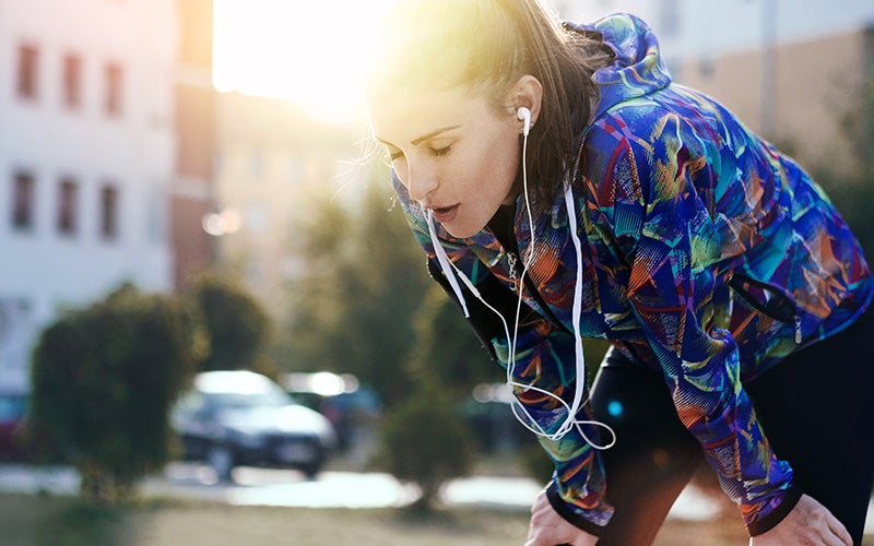 A white woman with white earbuds and a bright blue jacket leans over onto her knees in the middle of a strenuous workout outdoors