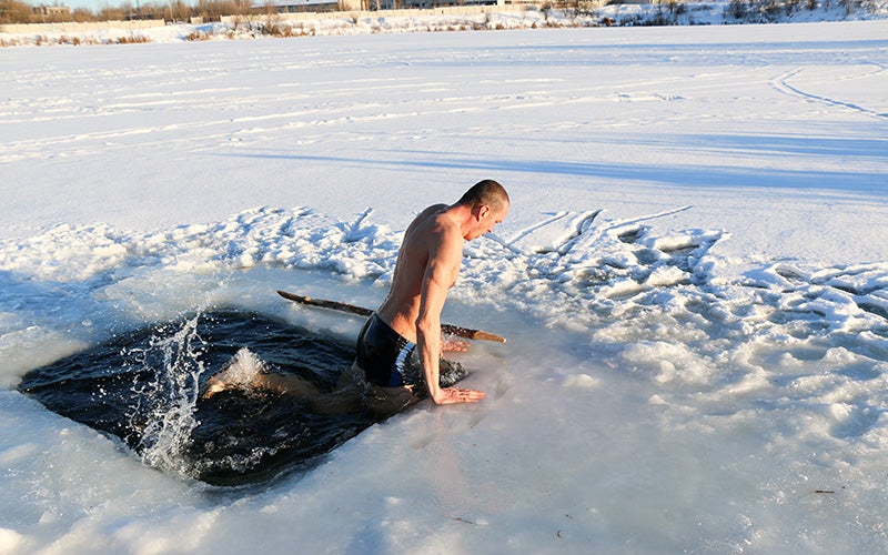 A white man pulls himself out of a hole in lake ice after taking an icy plunge for cold thermogenesis