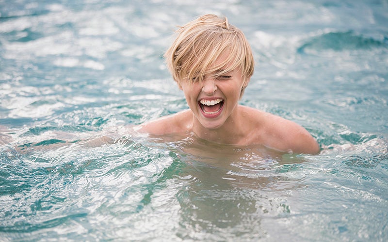 A grinning, topless model with a long blonde pixie cut shows an open-mouthed laugh while submerged to the shoulders in gently lapping water