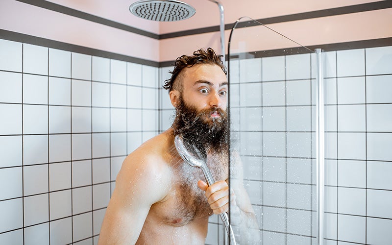 A white man with brown hair and a long brown beard stands topless in a glass-walled shower. He holds the shower head to his right shoulder. His eyes are spread wide in an expression of shock as if he has just been hit by the first jets of cold water.