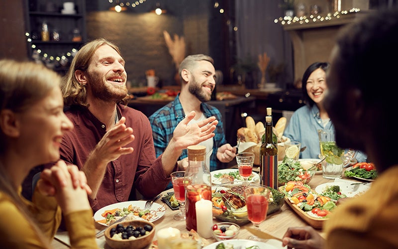 A group of friends - two white men, a white woman, and two people of color at the edges of the frame, laugh over a dinner table filled with salads, tapas, and a pitcher of sangria. The two white men's faces are centered and in focus in the photo.
