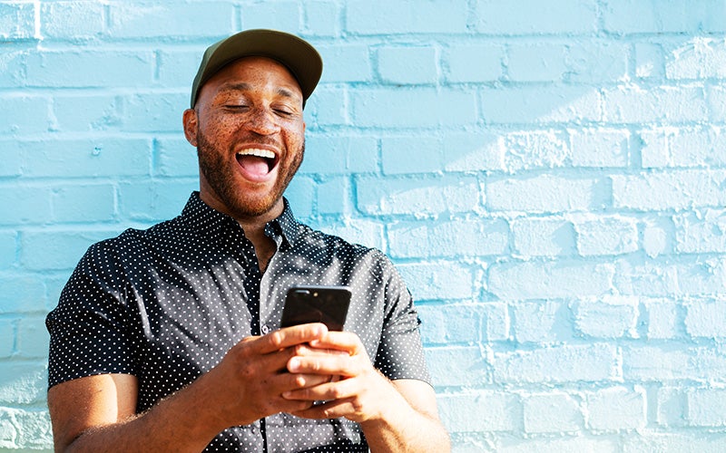 A man of color wearing a black baseball cap smiles, releasing dopamine and other happy hormones, while looking at his phone screen