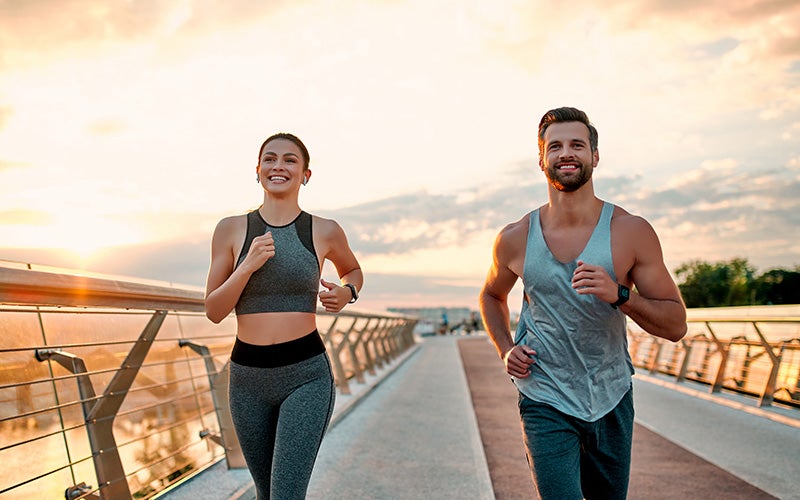 A white man and a white woman jog on a bridge outdoors in the light of the sunset. Both are smiling.