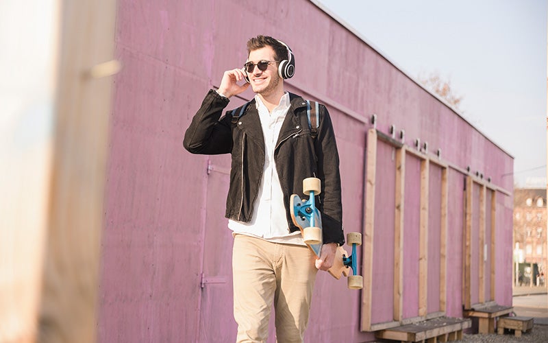 A white man in chinos, a white polo shirt with popped collar, and a short black zippered jacket carries a skateboard in one hand while the other holds his over-ear wireless headphones. He smiles and walks past a building painted pink.