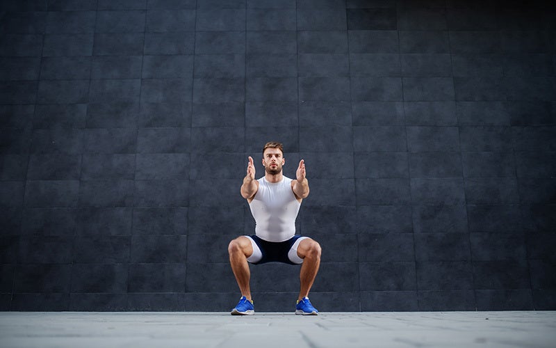 a white man doing an air squat with his own body weight in front of a gray marbled wall
