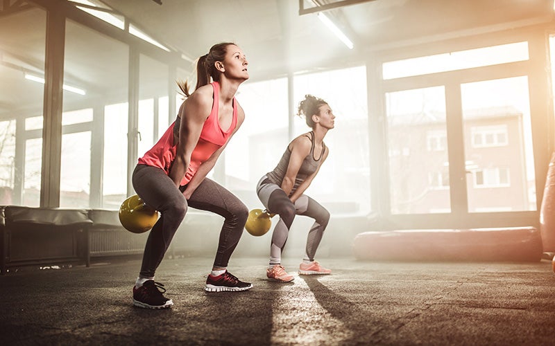 Two white women in tank tops stand in a gym, swinging kettlebells 