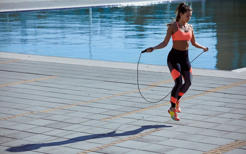 A medium-skin-toned woman in a pink sports bra and black and pink leggings jumps rope outdoors near a pool