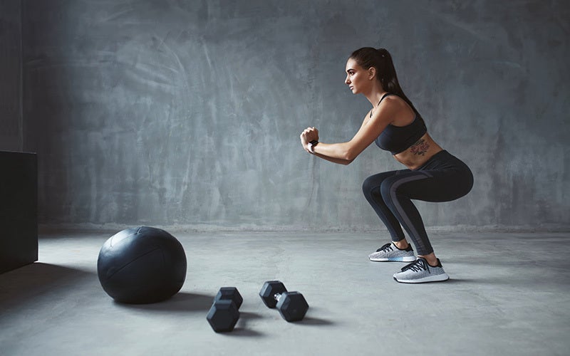 A white woman does squats with a weighted ball and two dumbbells in front of her.
