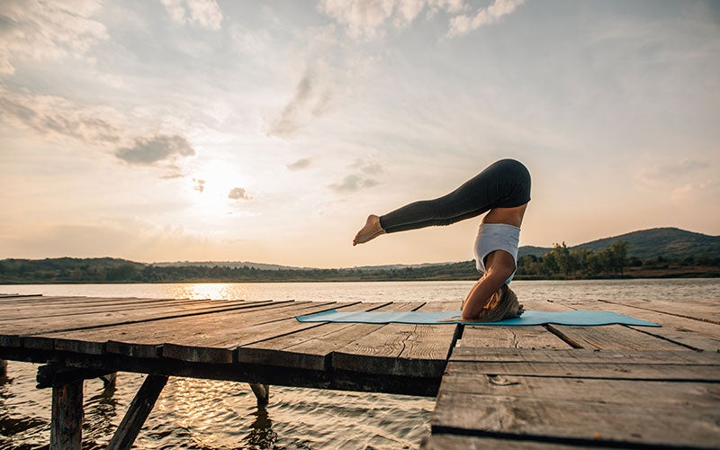 A white woman does a headstand on a pier slightly above water level. Her body is bent at the waist, with her legs stretched straight out at a 90-degree angle from her torso. She supports herself on her forearms, elbows, and head.