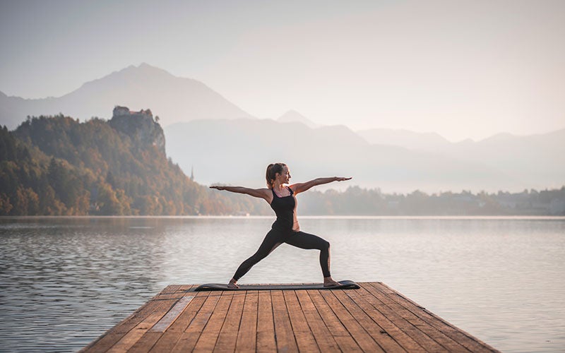 A white woman performs the warrior pose on a wooden pier with a lake and hills behind her.