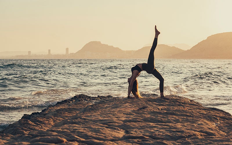 A white woman does a backbend with her right leg extended straight into the air on a rocky promontory with the waves lapping nearby.
