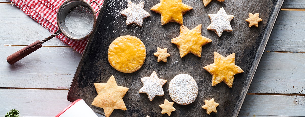 A well-used metal cookie sheet holds a batch of marzipan cookies. Some are dusted with powdered sugar, some are washed with egg yolk, and some are left plain. A sugar sifter sits next to the cookie sheet with leftover powdered sugar inside it.