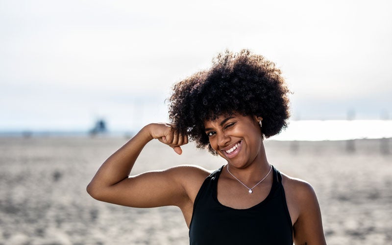 A woman of color with natural tightly curled hair flexes her biceps and winks at the camera with a grin. She is standing on a beach. 