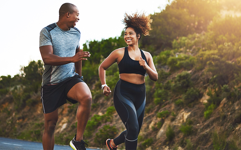 A man and woman of color grin at each other while taking a jog on an outdoor, paved road. A hill covered in greenery is visible to the woman's left side.