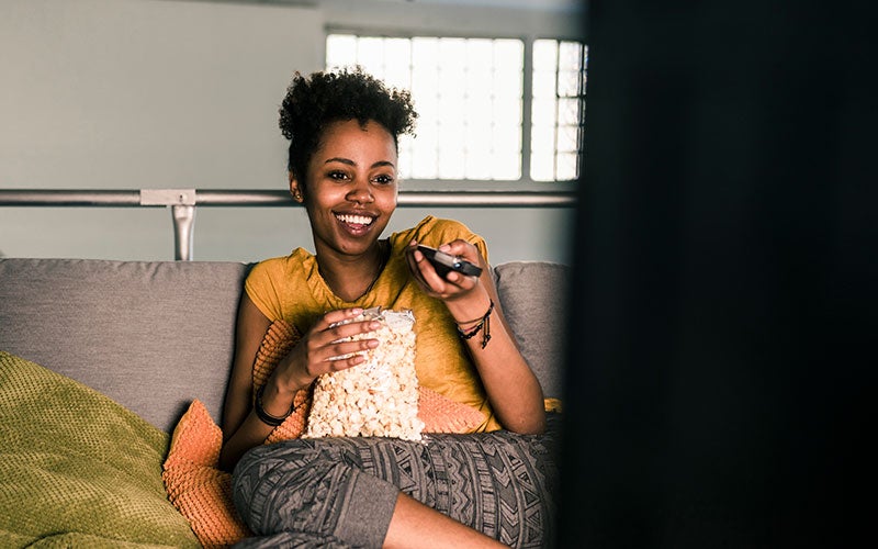 A woman of color smiles while holding a bag of popcorn in one hand, a remote in her other, using procrastination by watching something on television.