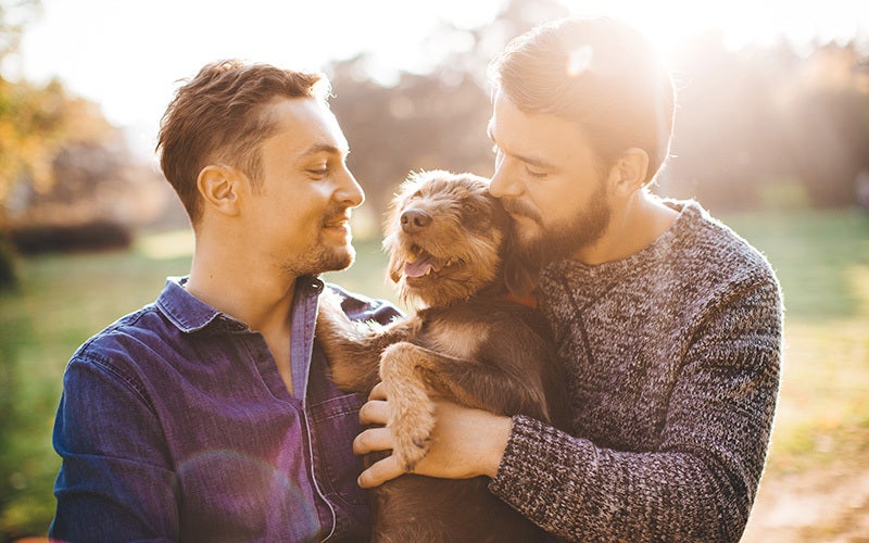 a white couple with beards and short hair snuggle with a dog to release happy hormones. They are standing outdoors with bright sunlight shining from behind the taller man's head.