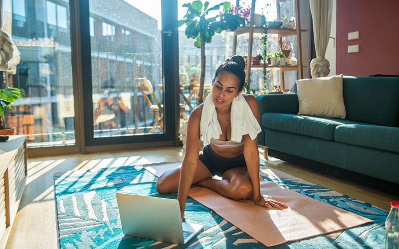 A woman of color sits on a yoga mat in a sunlit living room with a towel wrapped around her neck, with her laptop open to do a home workout