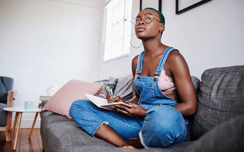 A Black woman wearing cat-eye glasses with teal-dyed close-cropped hair sits in overalls and a bra on a gray couch, holding a journal and pen, staring into the distance, wondering how to journal