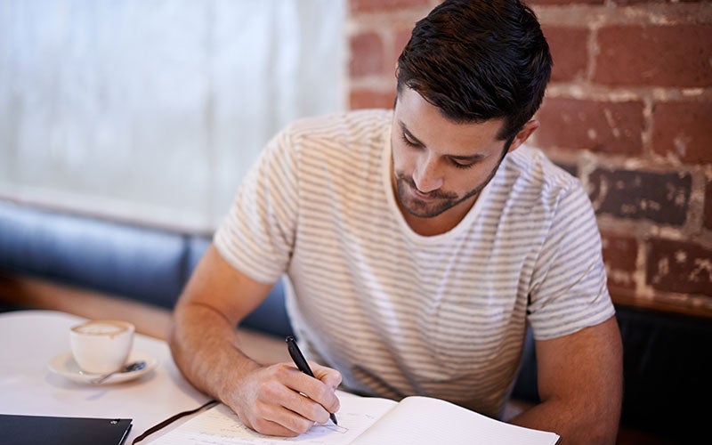 A white man with short, dark-brown hair wearing a short-sleeved gray-and-white-striped shirt sits at a table and writes by hand in a journal. There is a brick wall behind him and a cappuccino at his elbow.