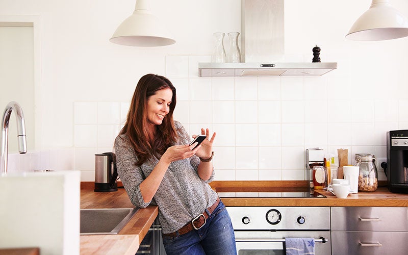 Eine Frau lehnt sich an den Küchentisch an und schaut dabei auf ihr Smartphone